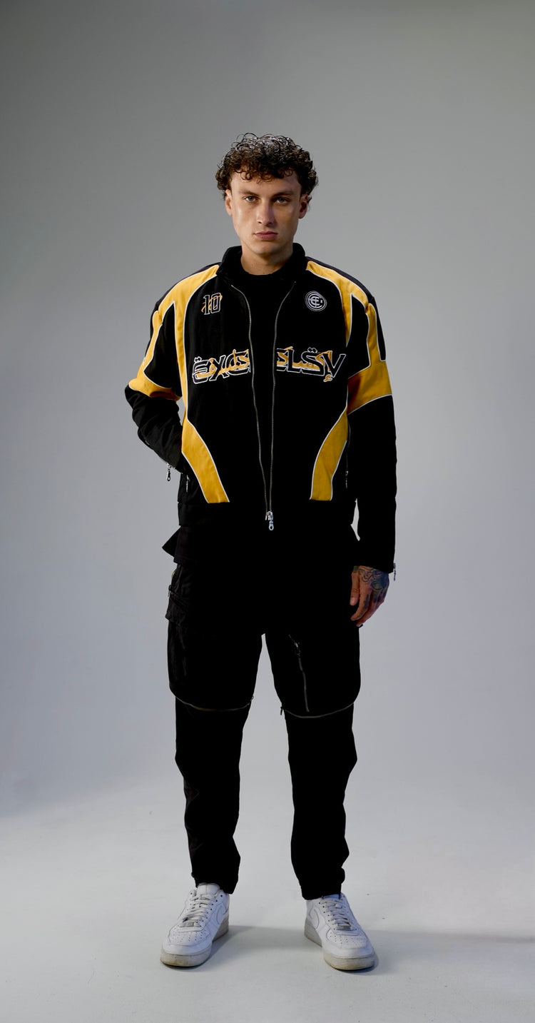A man modeling a black and yellow racing-style jacket and matching black pants against a light gray studio background.
